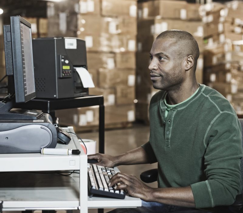 African American male warehouse worker working on a desktop computer in a large distribution warehouse ,with stacks of cardboard boxes in the background.