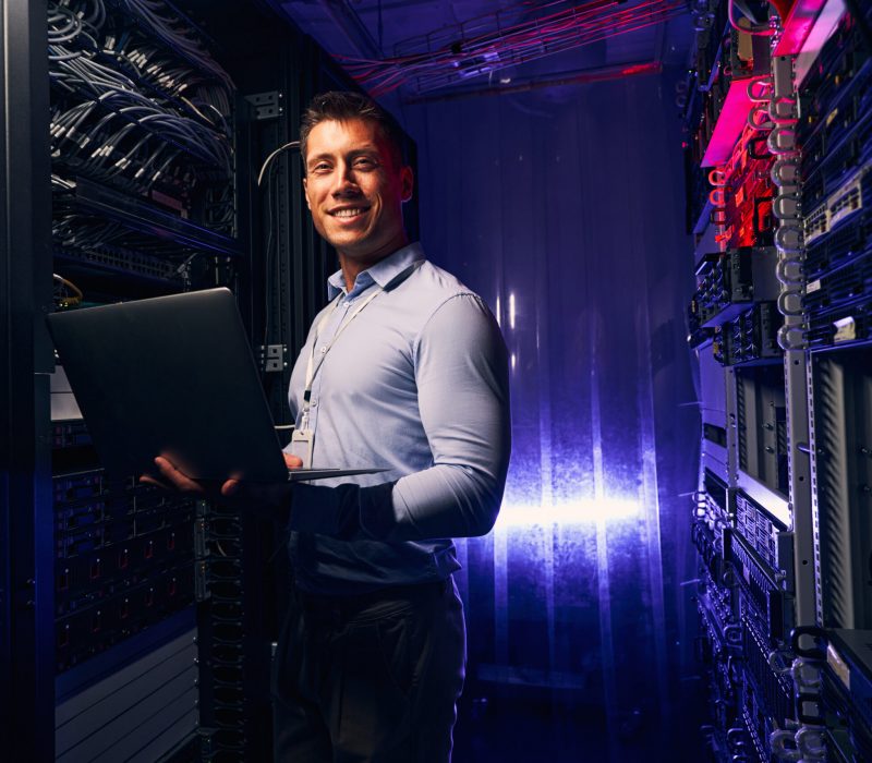 Smiling cheerful sysadmin with notebook computer in his hands standing beside open server rack cabinet